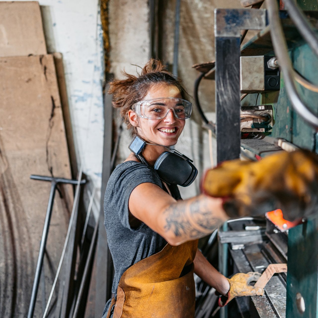 Eine Frau mit Schutzbrille und Schutzkleidung steht in einer Werkstatt und lächelt in die Kamera